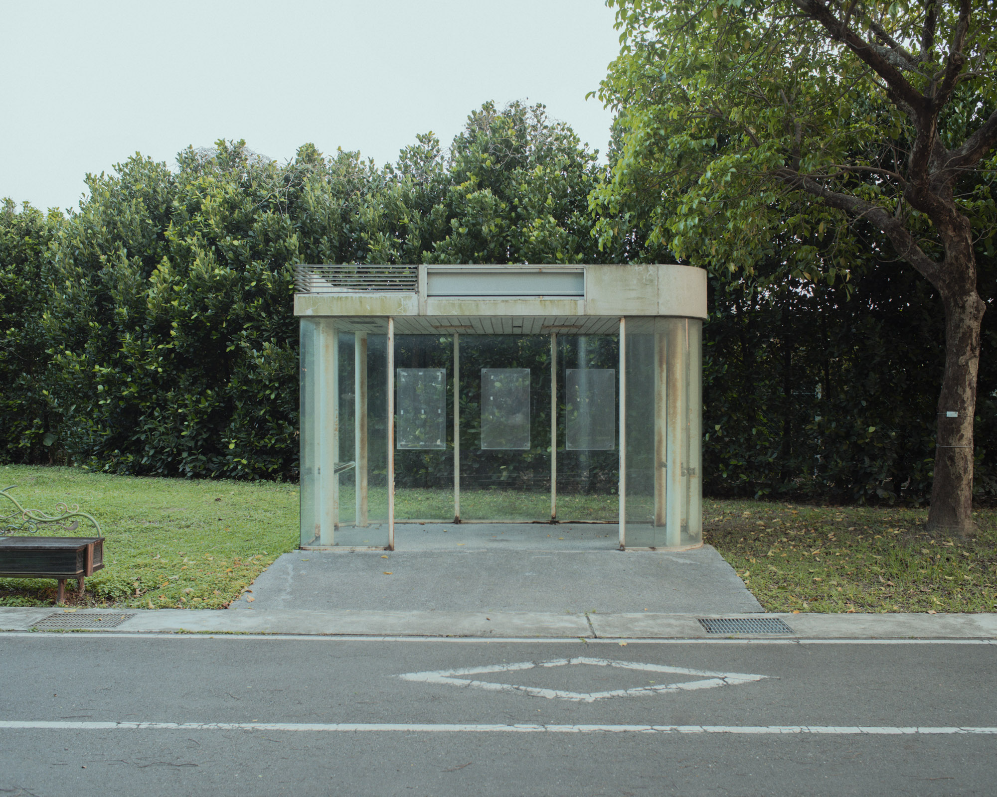 Gläsernes Bushäuschen vor einer grünen Hecke an einer leeren Straße